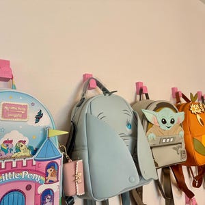 Puede incluir: Una colección de mochilas temáticas de personajes colgadas de ganchos rosas. Las mochilas presentan diseños de My Little Pony, Dumbo y Baby Yoda. Las bolsas son de varios colores, incluyendo azul, rosa, naranja y gris.