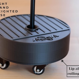May include: A black light stand weighted base with a circular design and a central opening for a pole. The base has a grooved top and a curved side, mounted on four rolling wheels. Text on the image reads: LIGHT STAND WEIGHTED BASE.