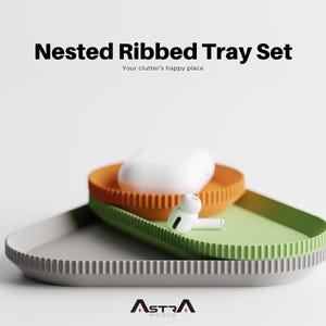 Puede incluir: Un juego de tres bandejas anidadas con bordes acanalados en gris, naranja y verde. Las bandejas están apiladas, con una funda blanca y un par de auriculares blancos encima. El texto "Nested Ribbed Tray Set" se muestra arriba.