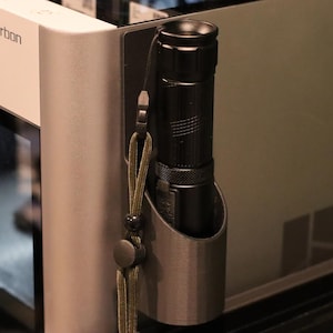 Puede incluir: Una linterna negra está asegurada en un soporte metálico fijado a un dispositivo. El soporte tiene un cordón con una borla decorativa. El dispositivo tiene el texto "X1-Carbon" impreso.