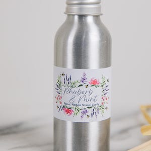 Puede incluir: Una botella de aromaterapia plateada con una etiqueta que dice "Rhubarb & Mint". La etiqueta presenta un diseño de corona floral con flores rosas y moradas y las palabras "Focus | Reduce Stress | Clarity."