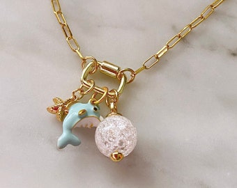 May include: A gold chain necklace with a blue dolphin charm, a small gold starfish charm, and a white crystal ball charm.
