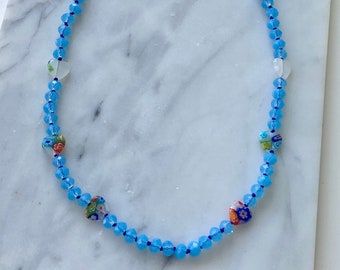 May include: A light blue crystal necklace with a gold chain clasp. The necklace features small, colorful heart-shaped beads with floral patterns.
