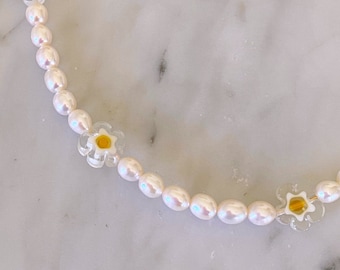 May include: A white pearl necklace with clear flower charms featuring yellow centers. The necklace is on a white marble surface.