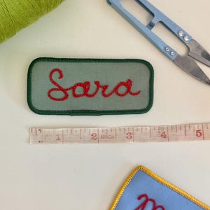 Pode incluir: Uma vista a&eacute;rea mostra um patch verde bordado com o nome "Sara" em fio vermelho. Tamb&eacute;m s&atilde;o vis&iacute;veis um par de tesouras azuis, uma fita m&eacute;trica e uma bobina de linha verde. Um patch azul com acabamento amarelo e bordado vermelho est&aacute; no canto inferior direito.