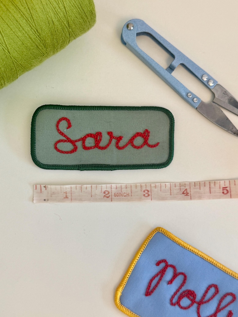 Pode incluir: Uma vista a&eacute;rea mostra um patch verde bordado com o nome "Sara" em fio vermelho. Tamb&eacute;m s&atilde;o vis&iacute;veis um par de tesouras azuis, uma fita m&eacute;trica e uma bobina de linha verde. Um patch azul com acabamento amarelo e bordado vermelho est&aacute; no canto inferior direito.