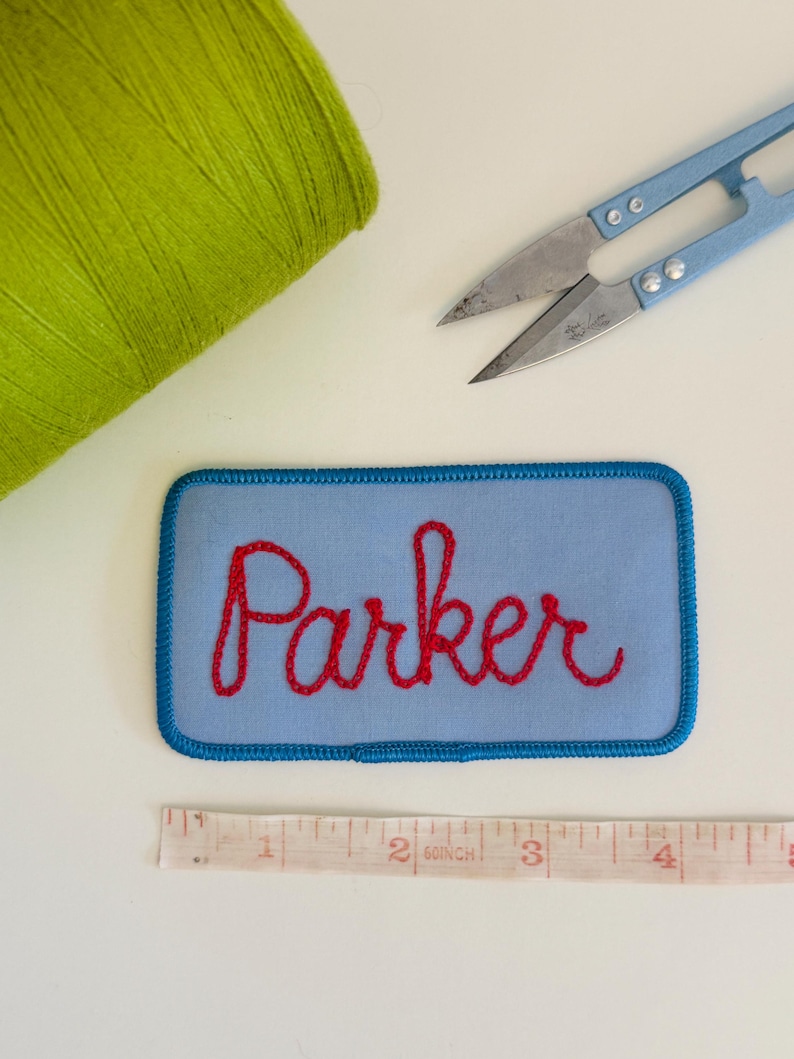 Pode incluir: Um patch bordado azul claro com o nome "Parker" em fio vermelho. O patch &eacute; retangular com cantos arredondados e uma borda azul. Uma bobina de linha verde e pequenas tesouras azuis tamb&eacute;m s&atilde;o vis&iacute;veis.