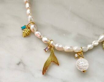 May include: A white pearl necklace with gold charms. The charms include a mermaid tail, a leaf, a crystal ball, and a small rectangular blue stone. The necklace is on a white marble surface.