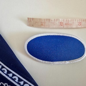 Pode incluir: Um patch oval azul com uma borda costurada branca. Uma fita m&eacute;trica de madeira est&aacute; ao lado do patch. Uma bandana azul marinho com um desenho paisley branco est&aacute; no canto.