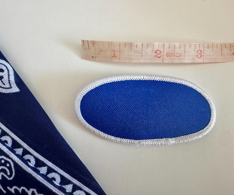 Pode incluir: Um patch oval azul com uma borda costurada branca. Uma fita m&eacute;trica de madeira est&aacute; ao lado do patch. Uma bandana azul marinho com um desenho paisley branco est&aacute; no canto.