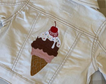May include: A white denim jacket featuring a large embroidered ice cream cone on the back. The design includes a tan waffle cone, a pink scoop, a brown scoop, white topping with colorful sprinkles, and a red cherry on top with visible stitching.