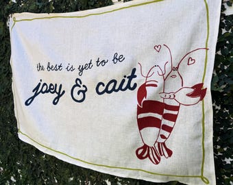 May include: Cream-colored banner with a green border and the words "the best is yet to be" above the names "joey & cait" in navy blue script. Two red lobsters are embracing, with heart accents.