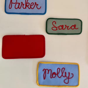 Pode incluir: Quatro patches de nome bordados. Os patches s&atilde;o retangulares com cantos arredondados. Os nomes "Parker" e "Molly" est&atilde;o em patches azul claro, "Sara" est&aacute; em um patch verde e um patch vermelho est&aacute; em branco. Os nomes est&atilde;o em it&aacute;lico vermelho.