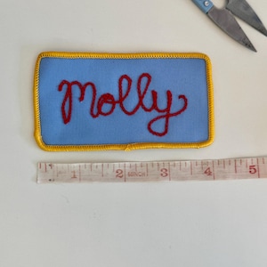 Pode incluir: Um patch retangular azul com o nome "Molly" bordado em fio vermelho. O patch tem uma borda dourada e mede aproximadamente 10 cm de comprimento. Uma tesoura azul est&aacute; no fundo.