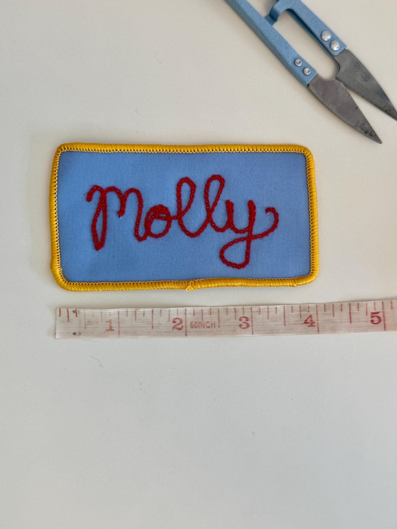 Pode incluir: Um patch retangular azul com o nome "Molly" bordado em fio vermelho. O patch tem uma borda dourada e mede aproximadamente 10 cm de comprimento. Uma tesoura azul est&aacute; no fundo.