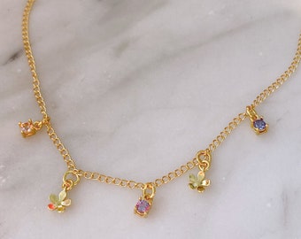 May include: A gold chain necklace with four charms: two small flowers, a purple gemstone, and a light blue gemstone.