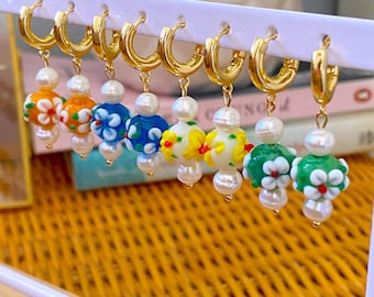 May include: A set of six pairs of gold hoop earrings with colorful glass flower charms and white pearl accents. Each charm features a different floral design with yellow, orange, blue, green, and white colors.