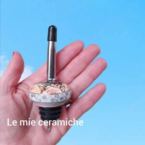 Può includere: Un versatore decorativo per bottiglie con punta nera e base in ceramica. La base in ceramica presenta un motivo floreale con accenti beige, verdi e rossi. Il testo "Le mie ceramiche" è visibile.