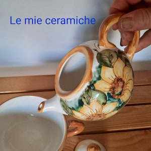 Può includere: Una teiera in ceramica con un designo a girasole. La teiera è bianca con accenti marroni e gialli. La teiera sta versando in una tazza in ceramica bianca.