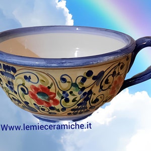 Può includere: Una grande ciotola in ceramica gialla con un manico blu. La ciotola è decorata con un motivo floreale in tonalità di blu, verde, rosso e giallo. La ciotola è su uno sfondo di cielo blu con nuvole bianche.