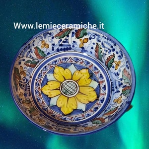 Op de afbeelding: Een blauwe en witte keramische kom met een geel bloemenmotief in het midden. De kom heeft een decoratieve rand met blauwe, witte en rode accenten. De kom staat op een blauwe en groene achtergrond.