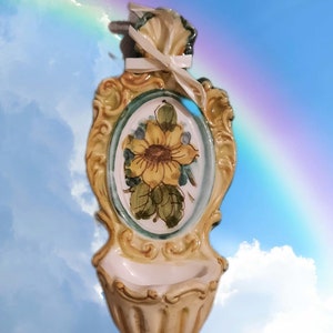 May include: Ceramic wall-mounted decorative piece with a sunflower design. The piece has a cream-colored base, a gold-colored frame, and a white ribbon. The text "Le mie ceramiche" is at the bottom.
