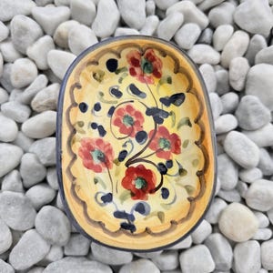Op de afbeelding: Ovale keramische schaal met een gele binnenkant, versierd met rode bloemmotieven en donkerblauwe accenten. De rand is geschulpt en heeft een bruine rand. De schaal staat op witte kiezelstenen.