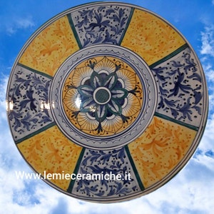 Può includere: Un grande piatto rotondo in ceramica con un design floreale blu, giallo e verde. Il piatto ha un centro rialzato con un design floreale più intricato. Il piatto è decorato con un bordo a festoni.
