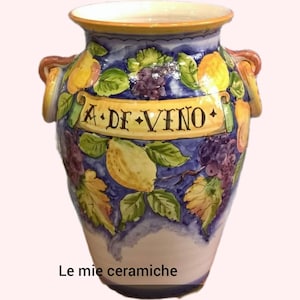 May include: A white ceramic pitcher with blue, yellow, and green painted fruit and grape designs. The pitcher has two handles and says "A. DE. VINO." on the front. The text "Le mie ceramiche" is written below the pitcher.