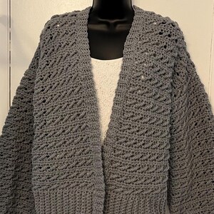 May include: A gray crocheted cardigan with a loose fit and a V-neck. The cardigan has a textured stitch pattern and long sleeves.