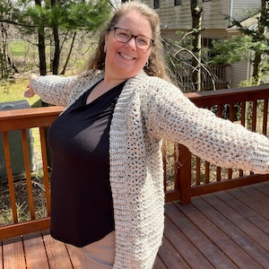 May include: A woman wearing a beige crocheted cardigan with a black shirt and khaki pants. She is standing on a wooden deck with a house and trees in the background.