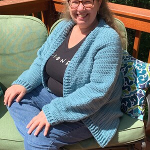 May include: A woman wearing a light blue crocheted cardigan with a black shirt that says "I.E.N.D." and blue jeans. She is sitting on a green chair on a wooden deck.