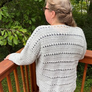 May include: A gray crocheted cardigan with a lacy pattern and a V-neck. The cardigan is worn by a person standing on a wooden deck.
