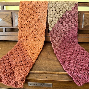 May include: Two rectangular crochet scarves featuring a textured granny stitch pattern. One scarf is a solid burnt orange color, while the other scarf is color-blocked with a diagonal transition from a light tan top to a deep maroon bottom section.
