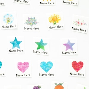 Puede incluir: Un conjunto de 24 etiquetas termoadhesivas de colores con varios diseños, incluyendo flores, estrellas, corazones y frutas. Cada etiqueta tiene el texto "Name Here" impreso en ella.