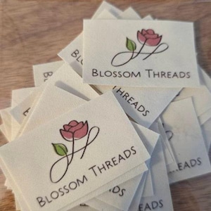 Puede incluir: Una pila de etiquetas de tela de color crema con una rosa rosa y el texto "Blossom Threads".