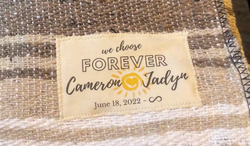 May include: A beige and brown woven blanket with a white label sewn on. The label has the text "we choose FOREVER Cameron Jadyn June 18, 2022 - ∞" with a yellow sun graphic and a heart.