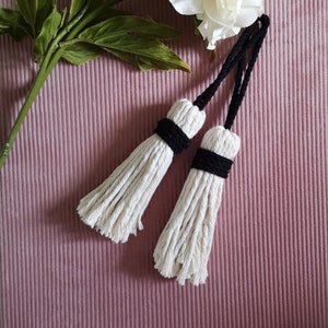 May include: Two white cotton tassels with black trim. The tassels are tied together with a black cord.