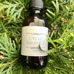 K&ouml;nnte beinhalten: Eine dunkelbraune Glasflasche mit schwarzer Pipette, beschriftet mit "CYCLE BALANCE" mit dem Text "TAKE 1-2 DROPPERS FILL WITH WATER DAILY" und "2oz|60ml". Die Flasche ist von gr&uuml;nem Laub umgeben.