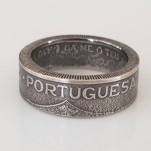 Könnte beinhalten: Ein silberner Ring mit dem Wort "PORTUGUESA" auf dem Band eingraviert. Der Ring hat ein strukturiertes, gealtertes Aussehen, mit zusätzlichem Text und Designelementen. Der Ring ist wahrscheinlich ein Schmuckstück.