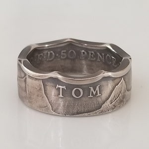 Puede incluir: Anillo de plata con el nombre "TOM" grabado. Hecho de una moneda de 50 peniques, con el texto "FD 50 PENCE" visible. El anillo tiene una apariencia texturizada y desgastada, mostrando el diseño original de la moneda.