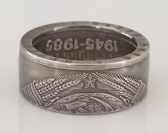 Könnte beinhalten: Ein silberner Ring mit detailliertem Design. Der Ring zeigt die Jahreszahlen "1945-1985" und ein Emblem mit Stern, Weizen und Landschaft. Der Ring hat ein breites Band und eine Vintage-Ästhetik.
