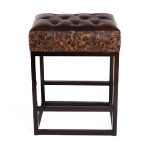 May include: A brown leather upholstered stool with a tufted top and a decorative floral pattern. The stool has a black metal frame with a rectangular base and horizontal supports. The stool is designed for seating.