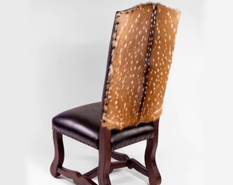 May include: A dark brown leather chair with a deer hide back. The chair has a wooden frame and is accented with small metal studs along the seat's edge. The deer hide features a pattern of brown spots on a tan background.