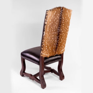 May include: A dark brown leather chair with a deer hide back. The chair has a wooden frame and is accented with small metal studs along the seat's edge. The deer hide features a pattern of brown spots on a tan background.