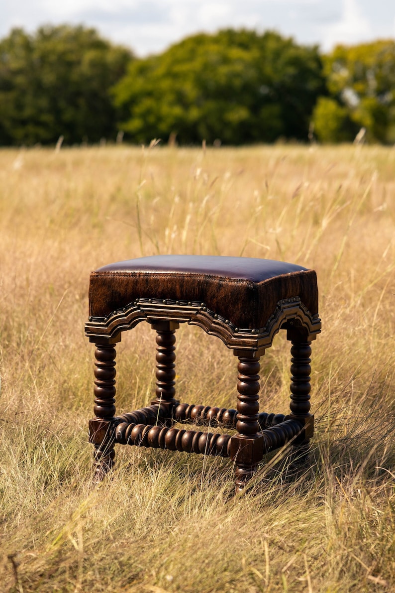 K&ouml;nnte beinhalten: Ein brauner Leder- und Holzhocker mit einem einzigartigen Design. Der Hocker hat eine rechteckige Sitzfl&auml;che mit einer braunen Lederplatte und einem dekorativen Holzrahmen. Die Beine sind aus Holz und haben ein gedrehtes Design.