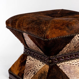 K&ouml;nnte beinhalten: Ein quadratischer Hocker mit einem braunen, schwarzen und wei&szlig;en Kuhfell- und Alligatorledermuster. Der Hocker hat eine gesteppte Oberseite und ist mit Metallnieten verziert.