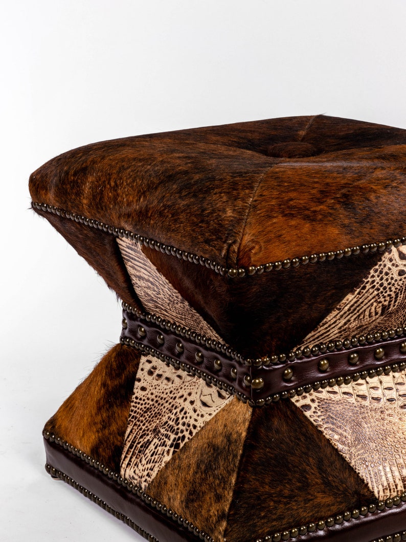 K&ouml;nnte beinhalten: Ein quadratischer Hocker mit einem braunen, schwarzen und wei&szlig;en Kuhfell- und Alligatorledermuster. Der Hocker hat eine gesteppte Oberseite und ist mit Metallnieten verziert.