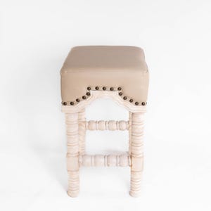 May include: A white wooden bar stool with a beige upholstered seat and decorative nailhead trim. The stool has a turned spindle design on the legs and a decorative arched back.