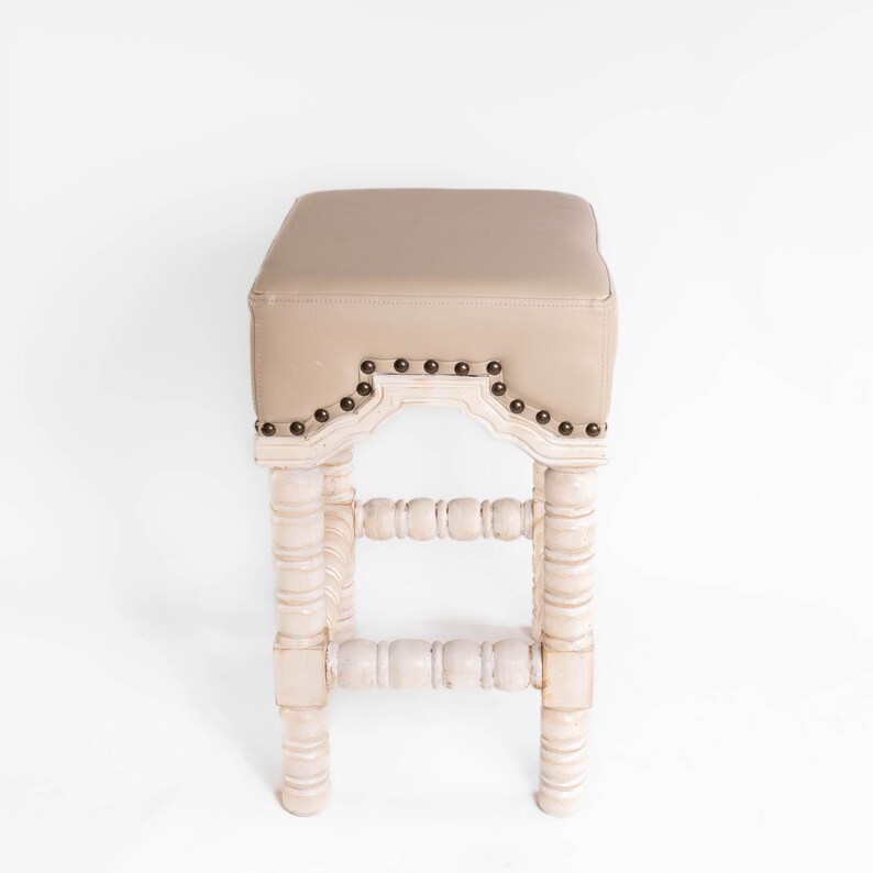 May include: A white wooden bar stool with a beige upholstered seat and decorative nailhead trim. The stool has a turned spindle design on the legs and a decorative arched back.